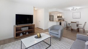 A modern living room with a flat-screen TV, wooden entertainment unit, grey armchair, dining area with a table and chairs, and a kitchen with white cabinets and appliances. - Landing | Chic 2BD, Courtyard (Paradise)