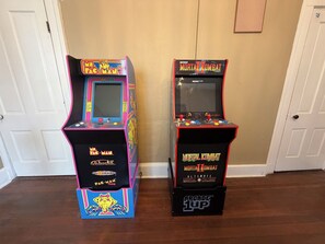 Two arcade machines, one labeled 'Pac-Man' and the other 'Mortal Kombat,' placed against a wall in a room with wooden flooring and white doors.