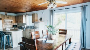 A kitchen with wooden cabinets, a refrigerator, a microwave, and a dining area with a table and chairs. - River & Miller Zoo | Louna-Nuage Shack (Frampton)