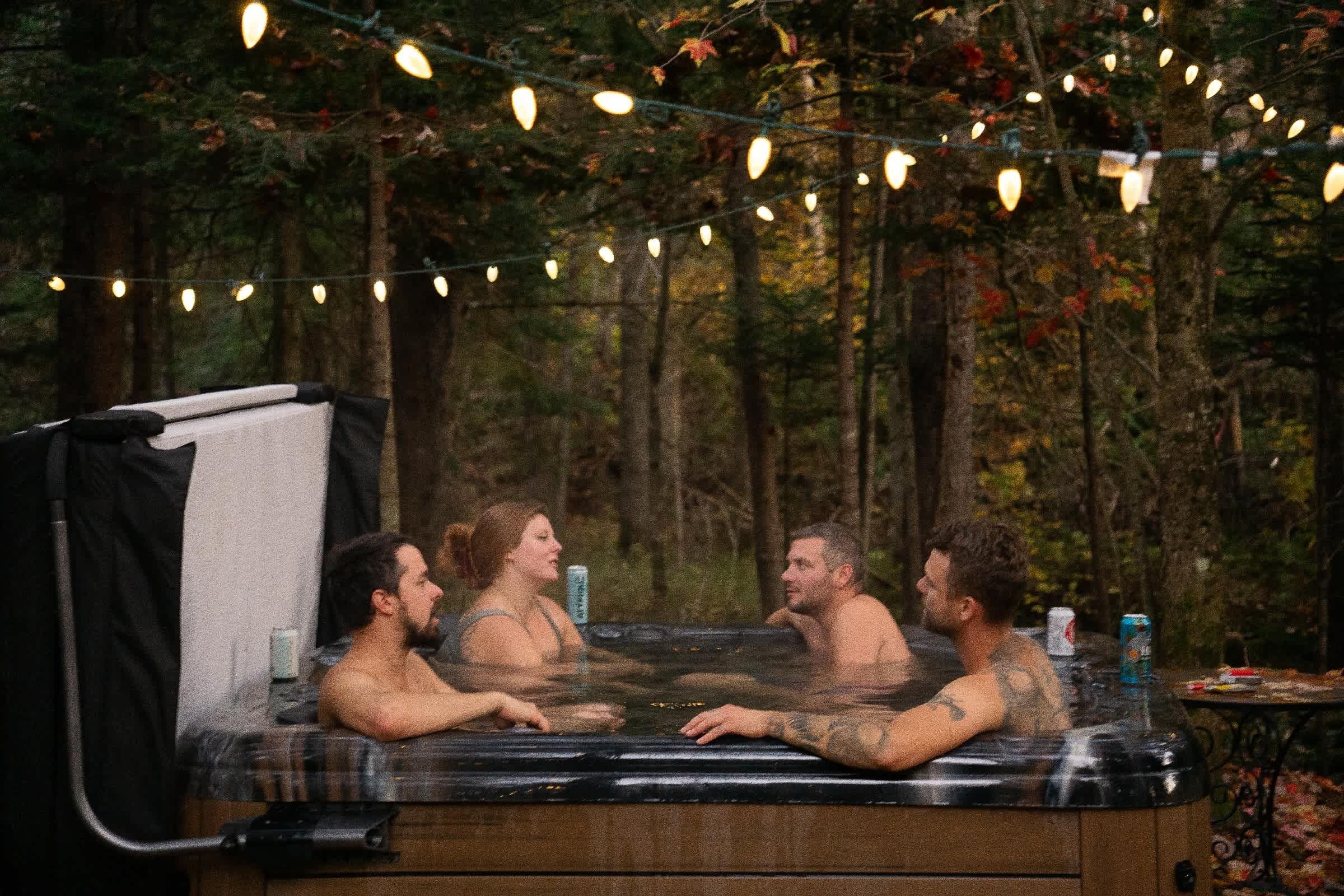 A hot tub with four people in a forested area, surrounded by string lights.