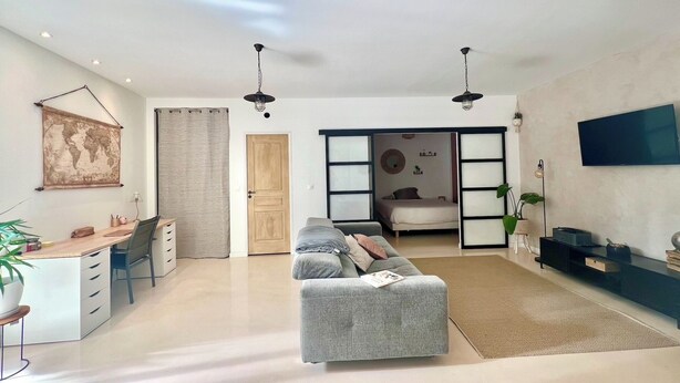 Una sala de estar moderna con un sofá gris, un escritorio con una silla, un televisor montado en la pared y una puerta corrediza que da a un dormitorio.