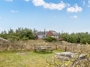 A house with a red roof, a chimney, and a wooden fence. There is a picnic table and a bench in the foreground.
