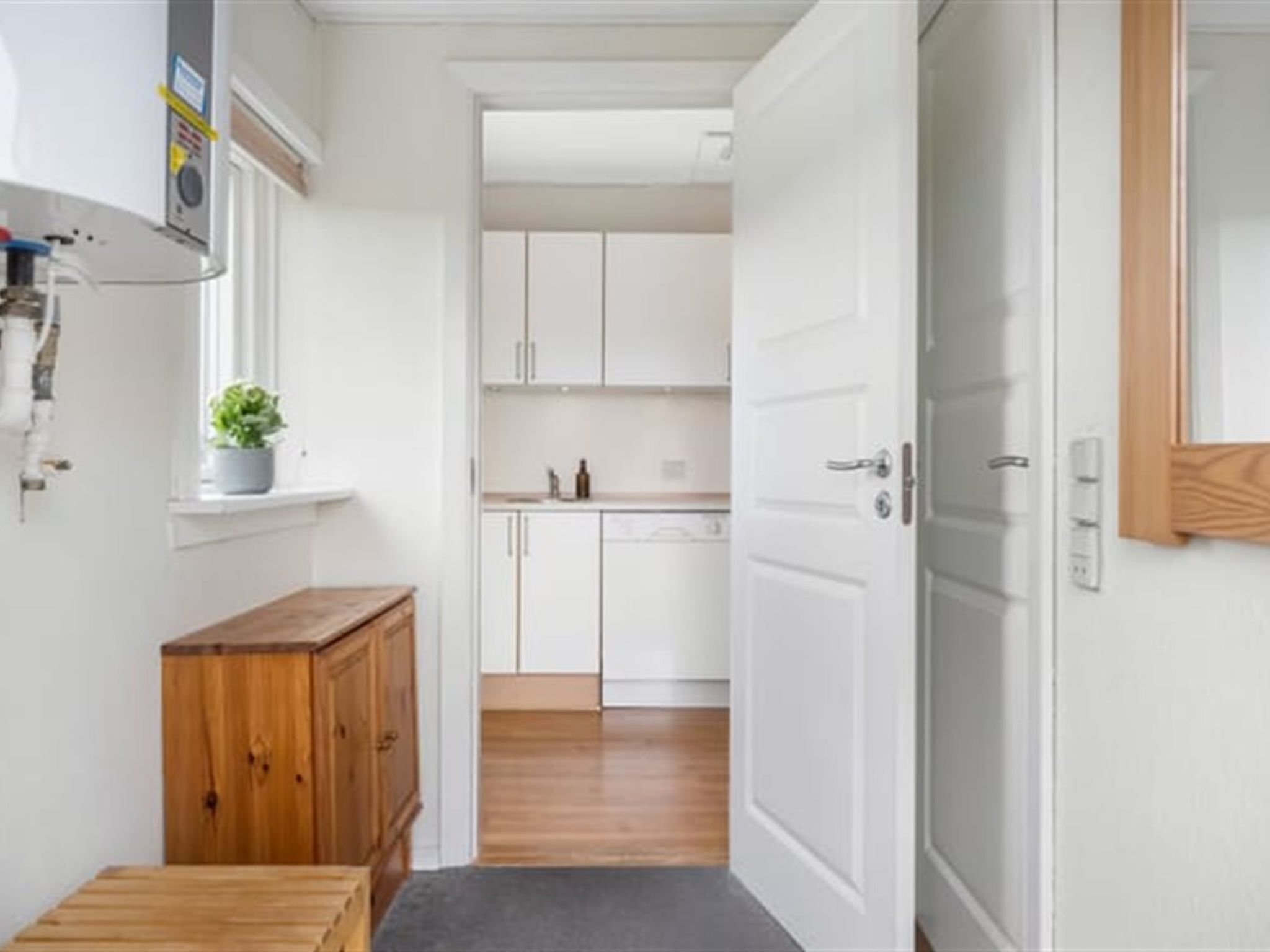A narrow hallway with a wooden bench, a potted plant, and white cabinets leading to a kitchen area.