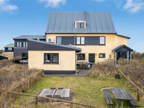 Une maison à deux étages avec un toit plat, un petit garage attenant et une cour avant agrémentée d’une table et de chaises en bois.