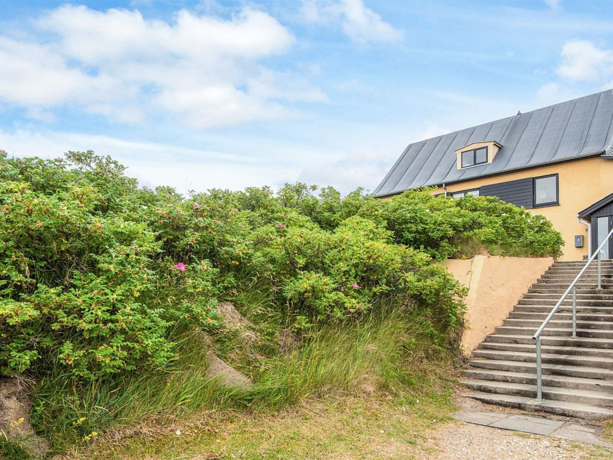 Ein Gebäude mit grauem Dach, einer Treppe, die zu einer Tür führt, und einem Garten mit Büschen und Blumen.