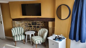 A room with a stone fireplace, two striped armchairs, a small round table, a white dresser with items on top, and a TV mounted on the wall. - Unicorn Inn (Deddington)