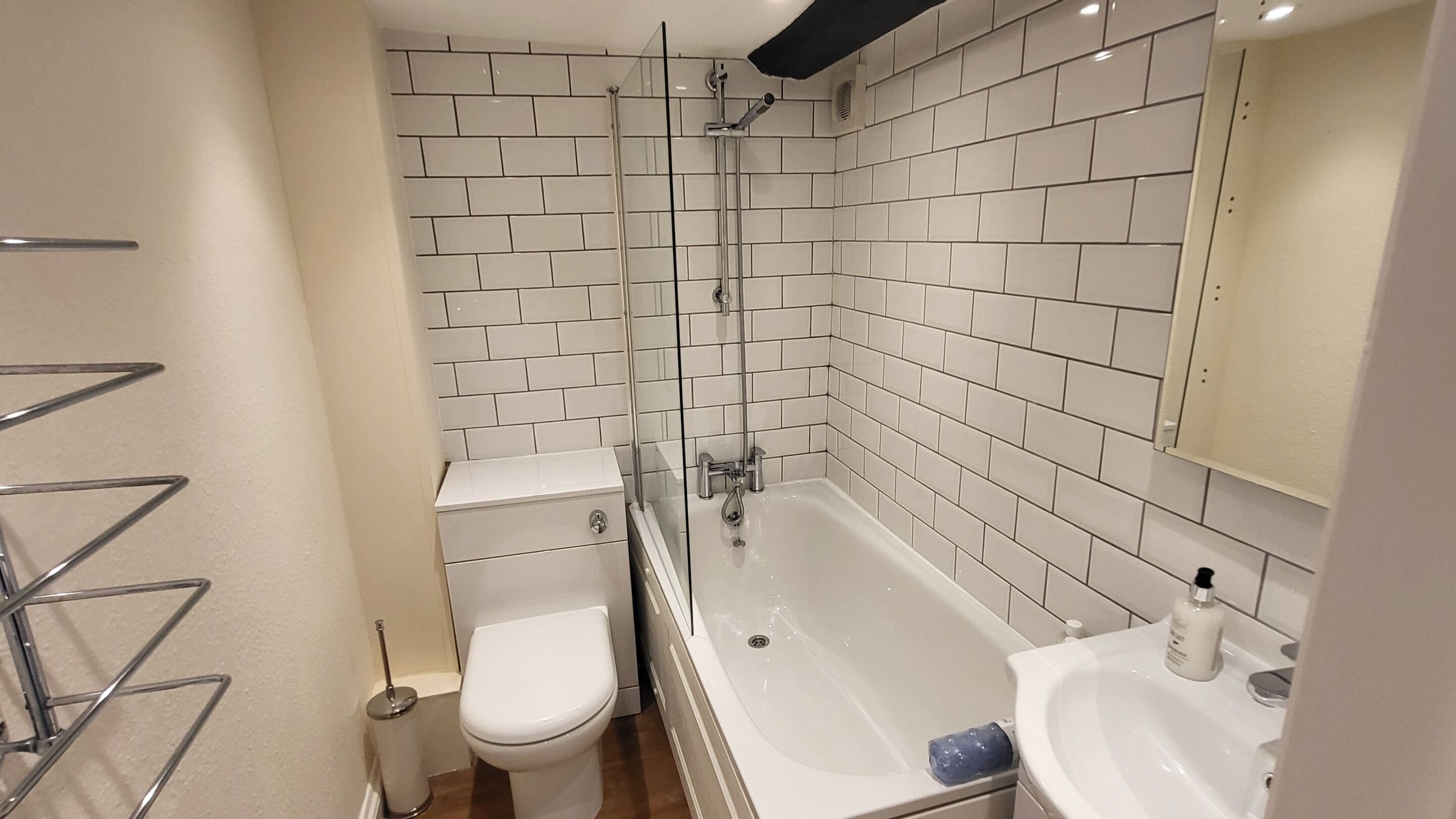 Un baño con paredes de azulejos blancos, cabina de ducha con mampara de vidrio, inodoro blanco, bañera y lavabo con espejo.