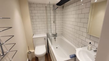 Un baño con paredes de azulejos blancos, cabina de ducha con mampara de vidrio, inodoro blanco, bañera y lavabo con espejo.