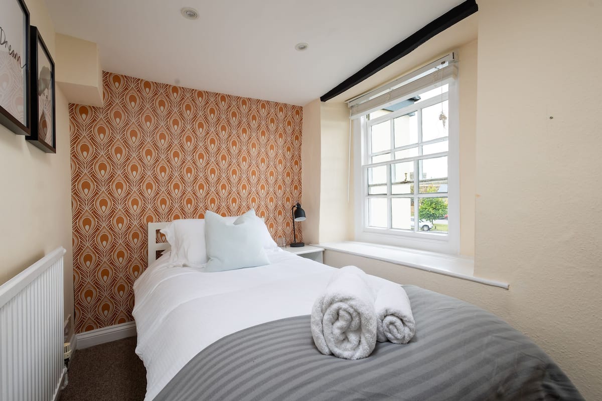 Une chambre d’hôtel avec un papier peint à motifs, un lit avec du linge de lit blanc, une fenêtre donnant sur un paysage et un radiateur.