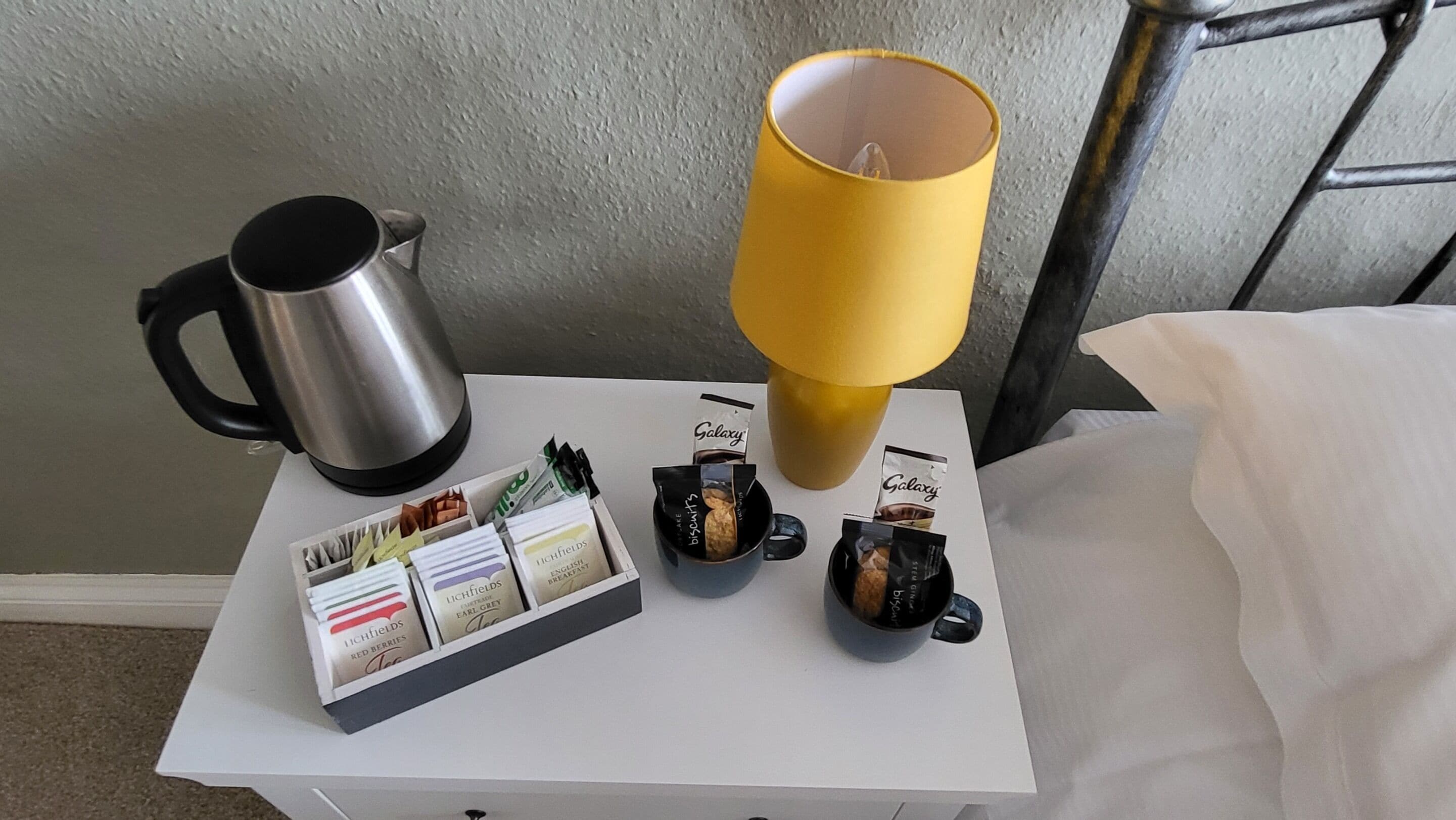 A white bedside table with a stainless steel kettle, a yellow lamp, and a box of Lichfields biscuits.