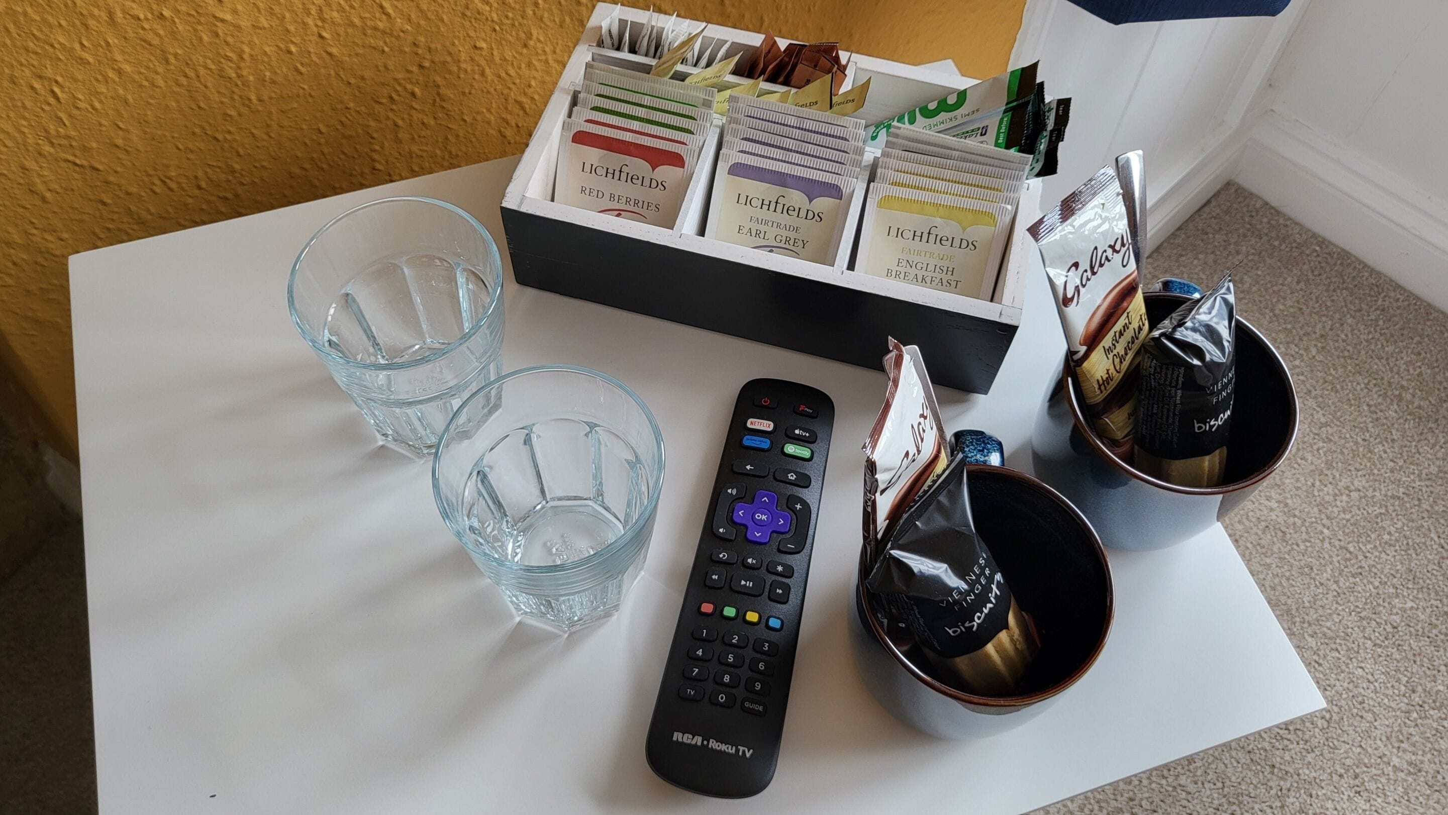 A table with a remote control, two glasses, a box of snacks, and two cups of instant coffee.