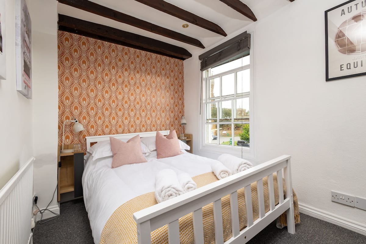 Une chambre à coucher avec un lit, des oreillers, une fenêtre et un mur recouvert d’un papier peint à motifs.