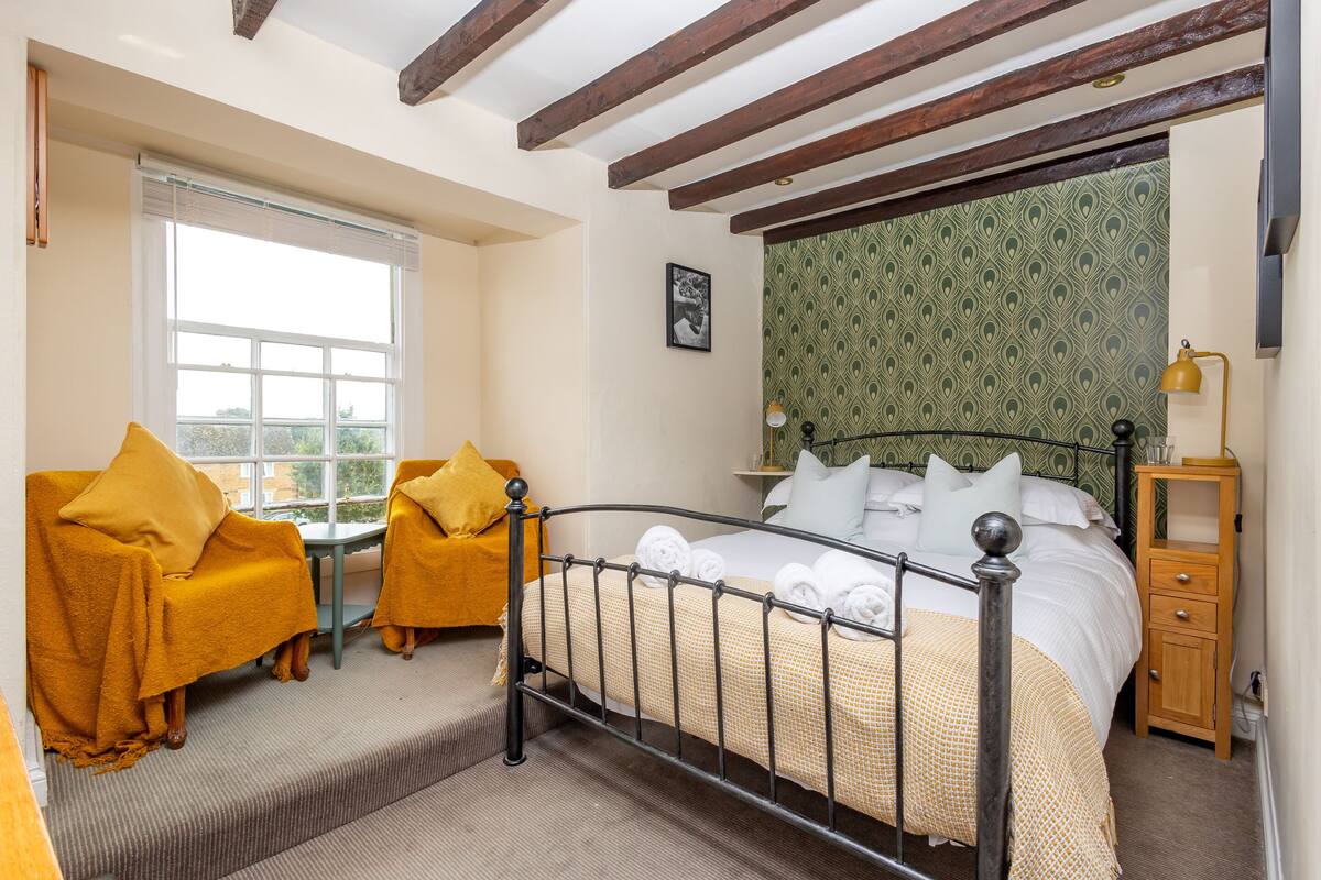 Une chambre d’hôtel comprenant un lit, deux fauteuils, une petite table, une table de chevet et une fenêtre avec des stores.