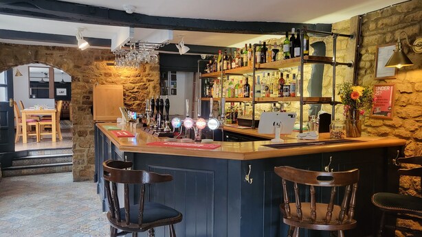 A bar area with a stone wall, wooden bar counter, and multiple liquor bottles. - Unicorn Inn (Deddington)