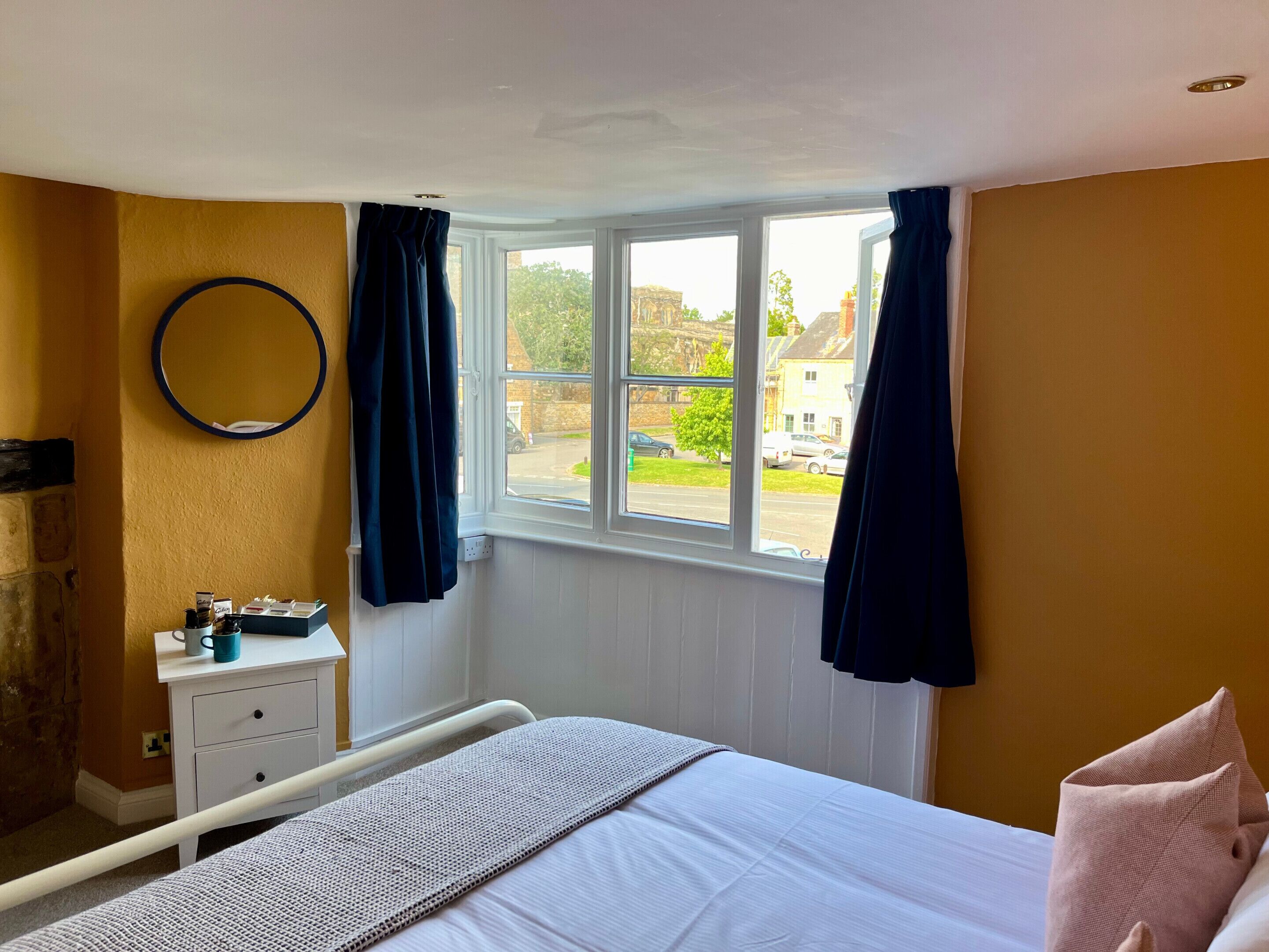 Quarto com cama, mesa de cabeceira, janela com cortinas e vista para a rua.