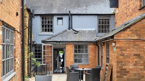 A narrow alleyway with a building featuring a grey facade, multiple windows, and a door. There are outdoor seating arrangements with chairs and tables. - Unicorn Inn (Deddington)