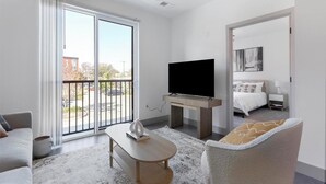 Ein modernes Wohnzimmer mit Flachbildfernseher, einer Couch, einem Couchtisch und einem Balkon mit Blick auf einen Parkplatz.