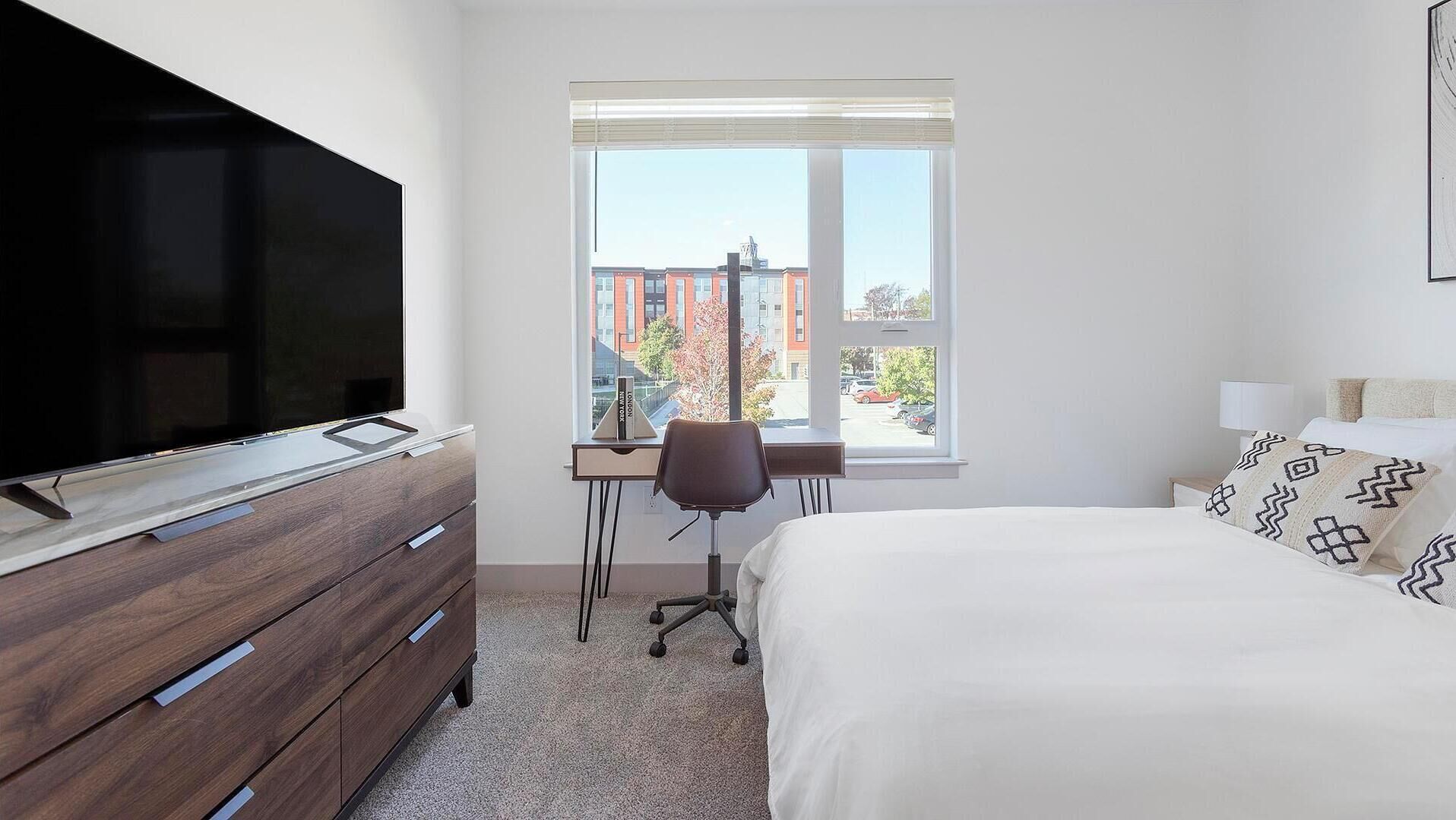 A hotel room with a bed, a desk, a chair, a television, and a window with a view of buildings.