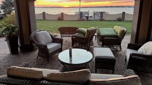 A patio with wicker furniture, a glass table, and a view of the sunset over the water. - High-End Waterfront Estate in Stratford | Relax, Unwind, Enjoy Views (Stratford)