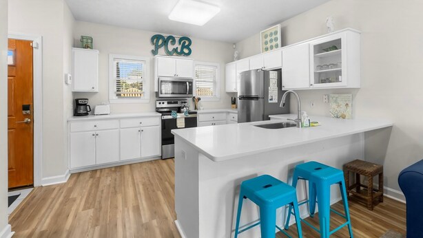A modern kitchen with white cabinetry, a central island, blue bar stools, and a 'PCB' sign on the wall. - Steps to Beach! Sleeps 8 + pool + garage+ beach gear + lagoon fishing + 2 patios (Panama City Beach)