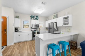 A modern kitchen with white cabinetry, a central island, blue bar stools, and a 'PCB' sign on the wall. - Steps to Beach! Modern Townhome w/ Pool, 2 Car Garage & Patios (Panama City Beach)