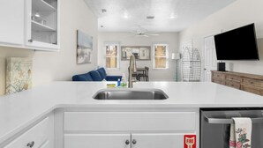 A modern kitchen with white cabinetry, a stainless steel sink, and a built-in oven. There is a dining area with a blue couch and a ceiling fan. - Steps to Beach! Sleeps 8 + pool + garage+ beach gear + lagoon fishing + 2 patios (Panama City Beach)