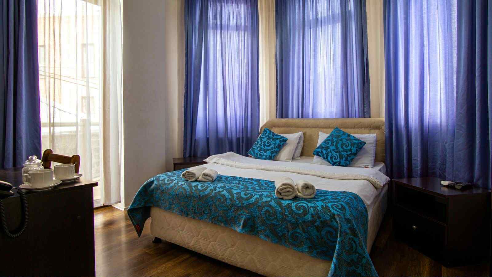 A hotel room with a bed, blue curtains, a wooden nightstand, and a small table with a telephone and cups.