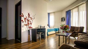 A living room with a sofa, a dining table, a chair, a bed with a blue cover, a TV on the wall, and a vase with flowers. - Hotel Nebula Inn (Tbilisi)