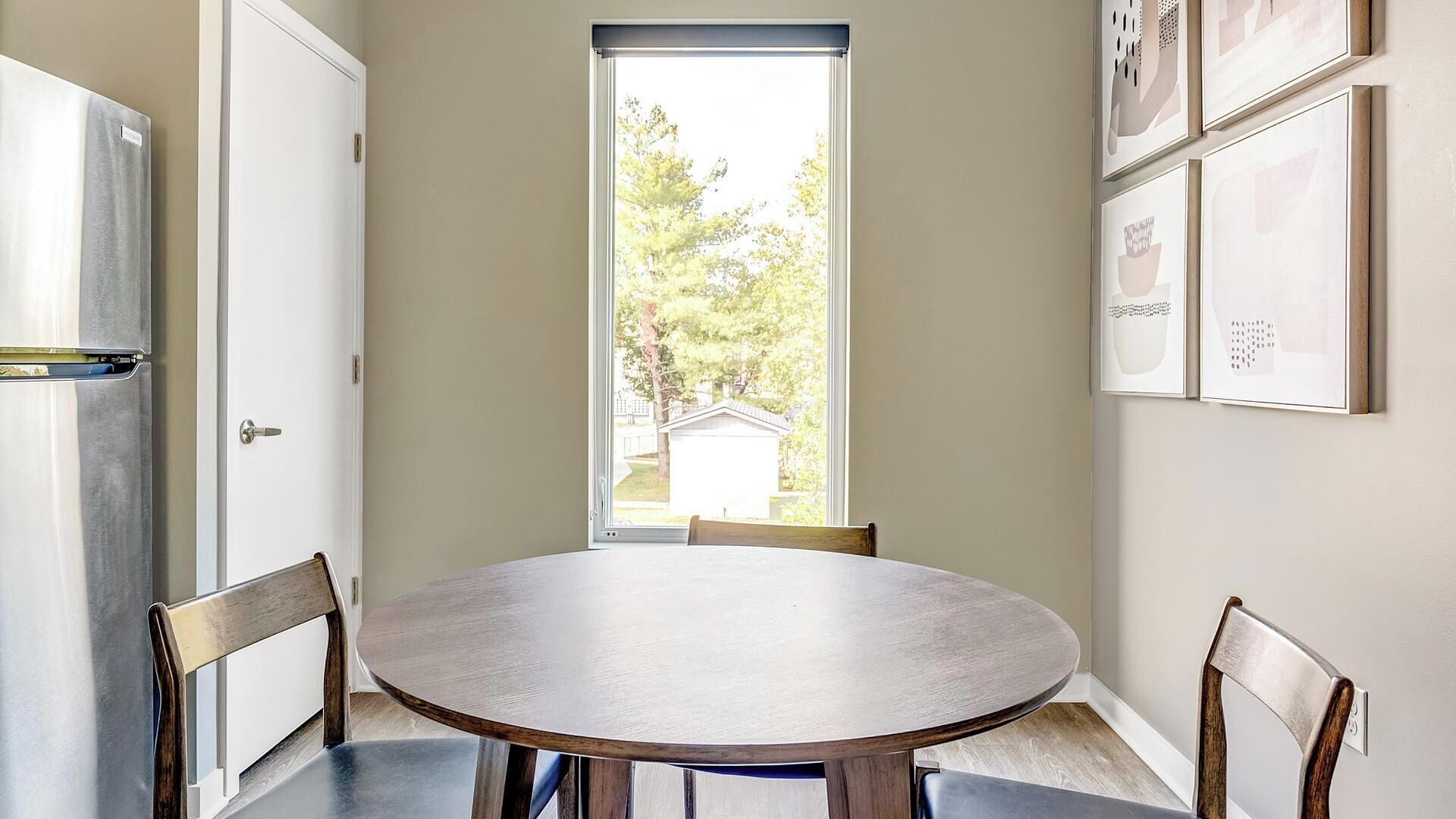 A dining area with a round table, chairs, a refrigerator, and a window with a view of trees and a shed.