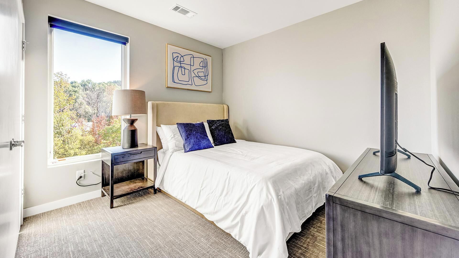 A bedroom with a bed, a bedside table with a lamp, a TV stand, and a window with a view of trees.