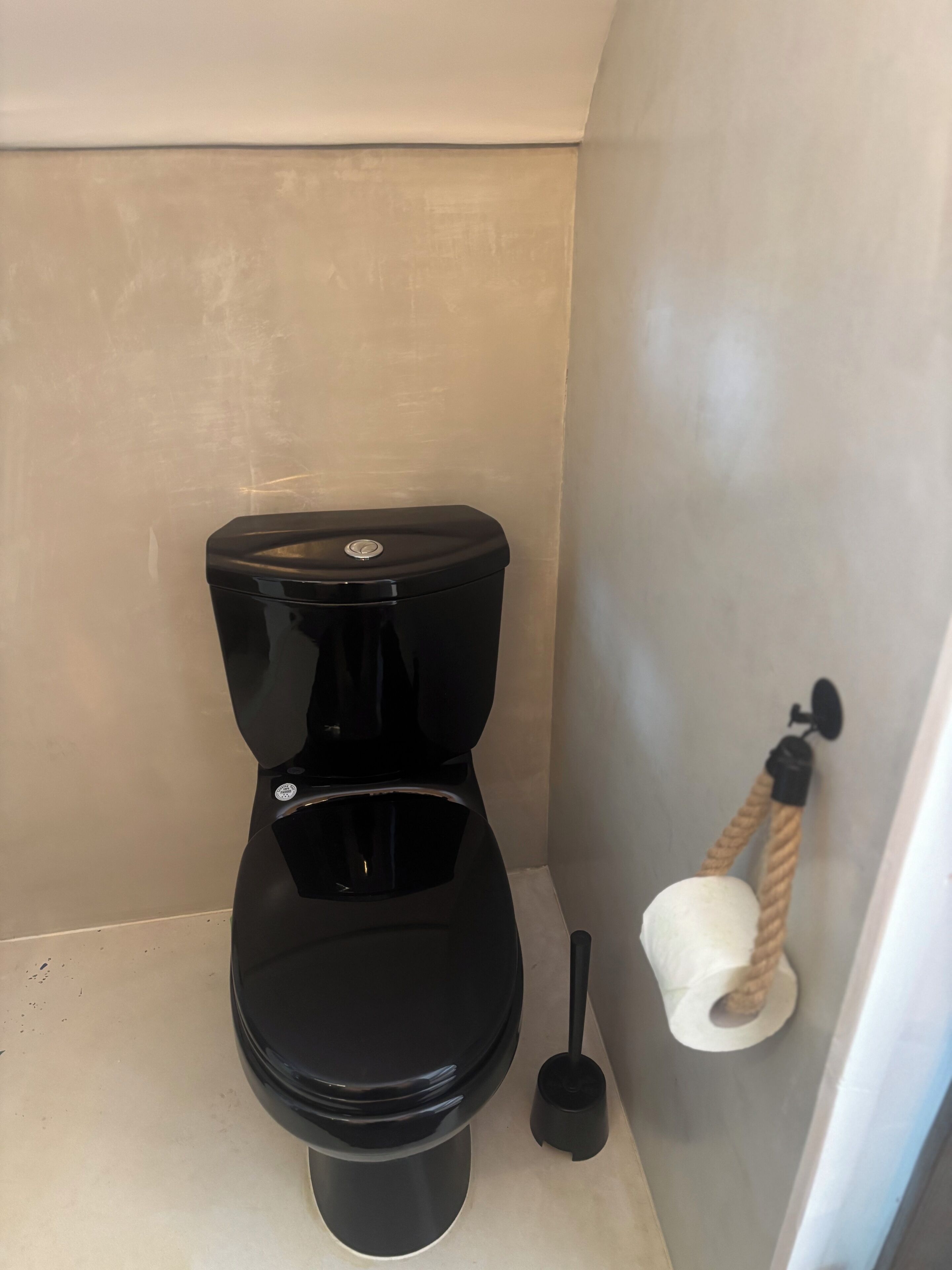Et toilet med sort låg og tank, en renskebakke og en toiletpapirholder med snor.
