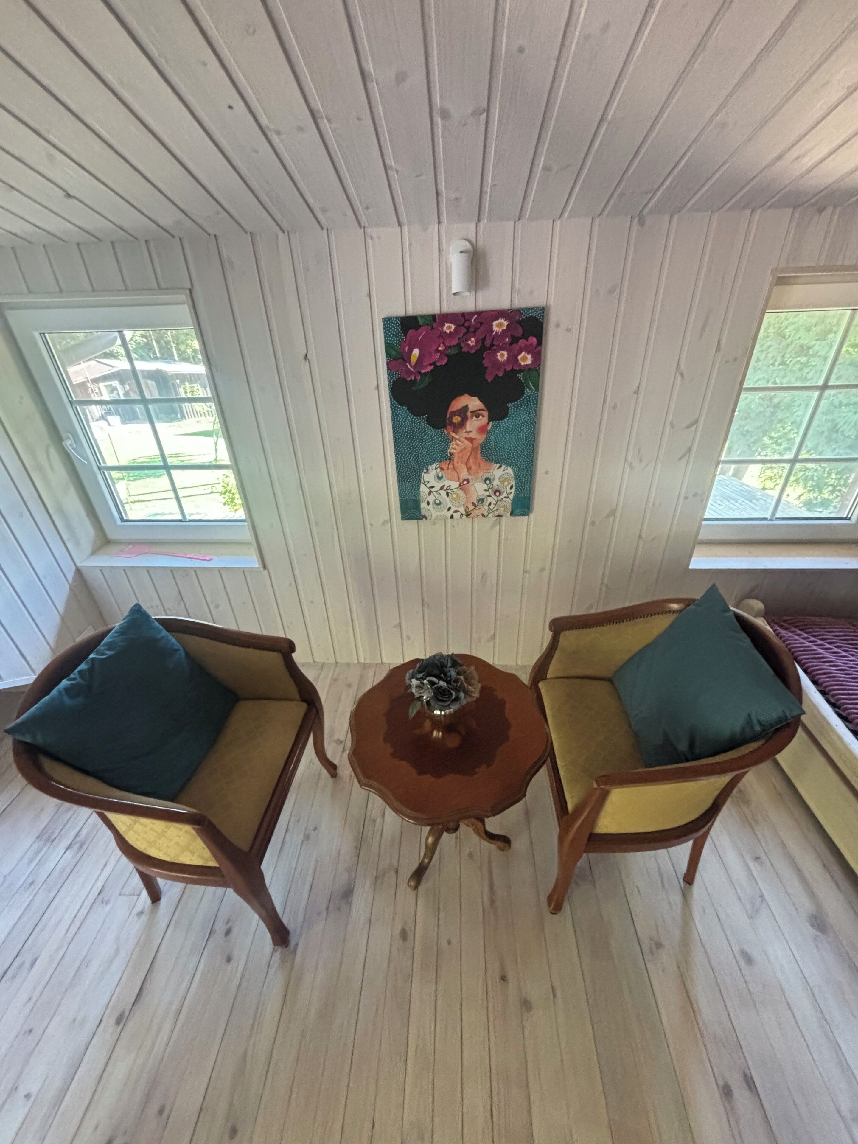 A room with wooden paneled walls, two wooden chairs with cushions, a small round table with a vase, and a painting on the wall.