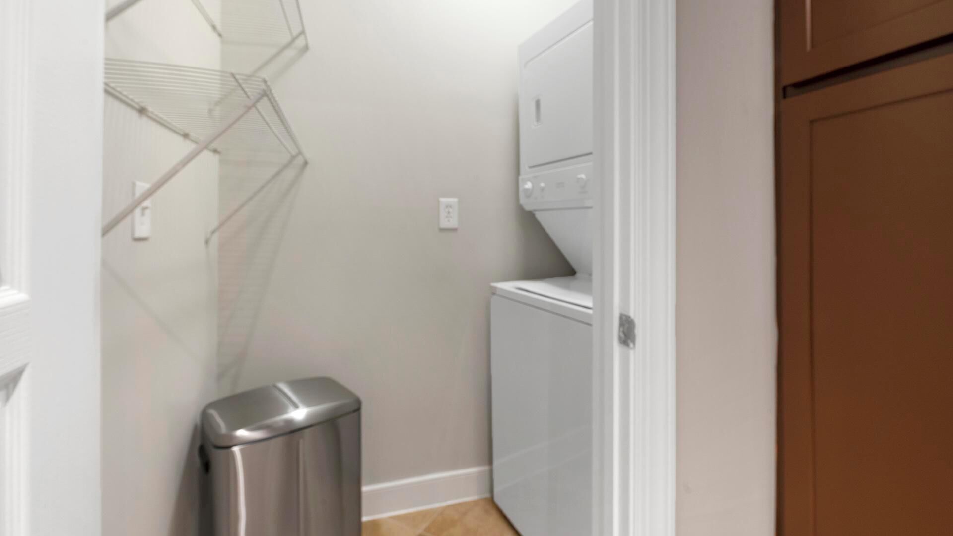 A laundry room with a washing machine, a metal trash can, and a wire shelf.