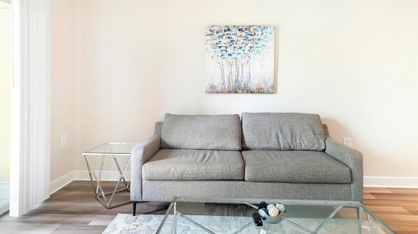 A living room with a grey sofa, a glass coffee table, and a painting on the wall. - Landing | Spectacular 1BD, Pool, Courtyard (Hyattsville)