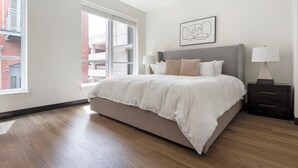 A bedroom with a large bed, a nightstand, a lamp, and a window with a view of buildings. - Landing | Beautiful 2BD, Courtyard, Gym (St. Louis)