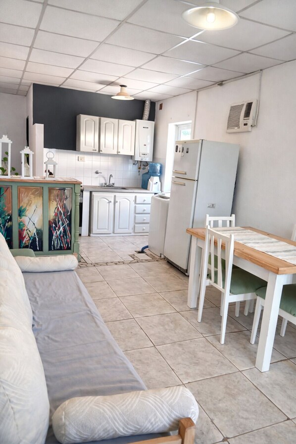 Une cuisine compacte avec des meubles blancs, un réfrigérateur, une table à manger avec des chaises et un canapé avec des coussins.