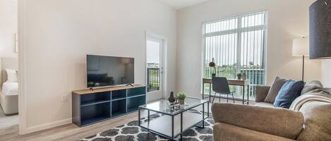 A modern living room with a flat-screen TV, a glass coffee table, a sofa, and a large window with a view.