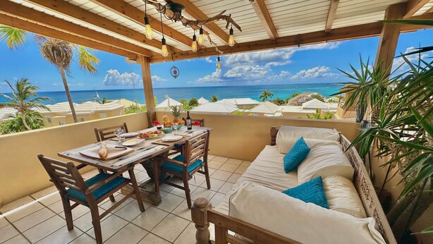 Eine Holzterrasse mit Frühstückstisch, einer Couch mit Kissen und Blick auf das Meer.