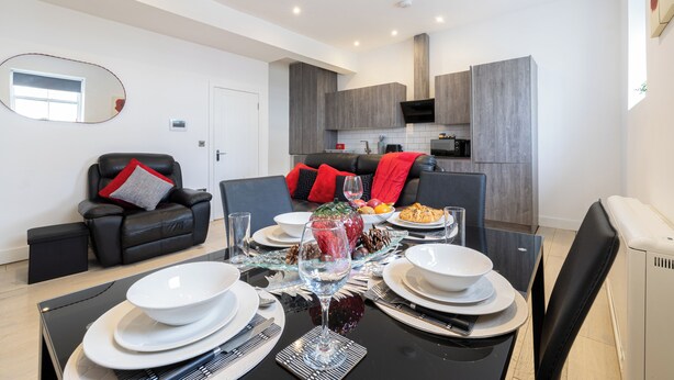 Een moderne woonkamer met een eettafel gedekt voor vier personen, een zwarte leren bank met rode kussens, en een keuken op de achtergrond.