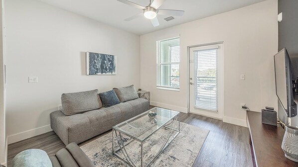 A living room with a sofa, a glass coffee table, a ceiling fan, and a door with blinds. - Landing | Modern 1BD, Gym, Pool (Tampa)