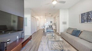 A modern living room with a flat-screen TV, a wooden entertainment unit, a glass coffee table, a grey sofa with blue cushions, and a kitchen in the background. - Landing | Modern 1BD, Gym, Pool (Tampa)