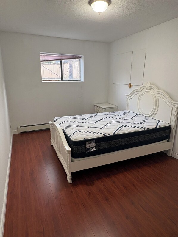 Une chambre avec un lit blanc, une petite table de chevet, une fenêtre et un radiateur.