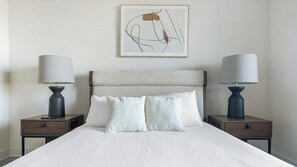 Habitación de hotel con una cama, dos mesitas de noche, dos lámparas, una obra de arte abstracta enmarcada y un aparador con cajón.