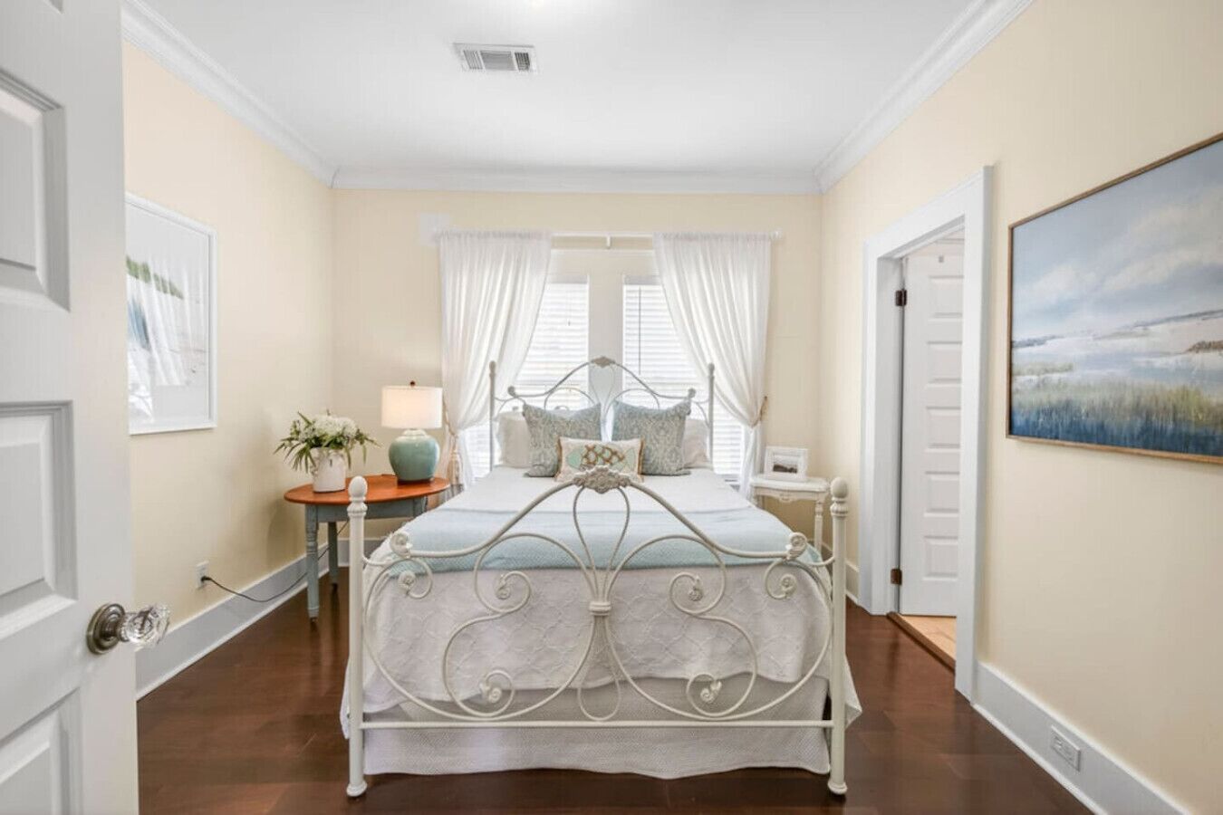 A bedroom with a metal bed frame, a bedside table with a lamp, a window with curtains, and a painting on the wall.