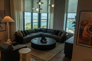 A modern living room with a sectional sofa, a round coffee table, and large windows offering a view of a bridge and water. - Skyview Studio on 34th | City & Bay Views (Miami)