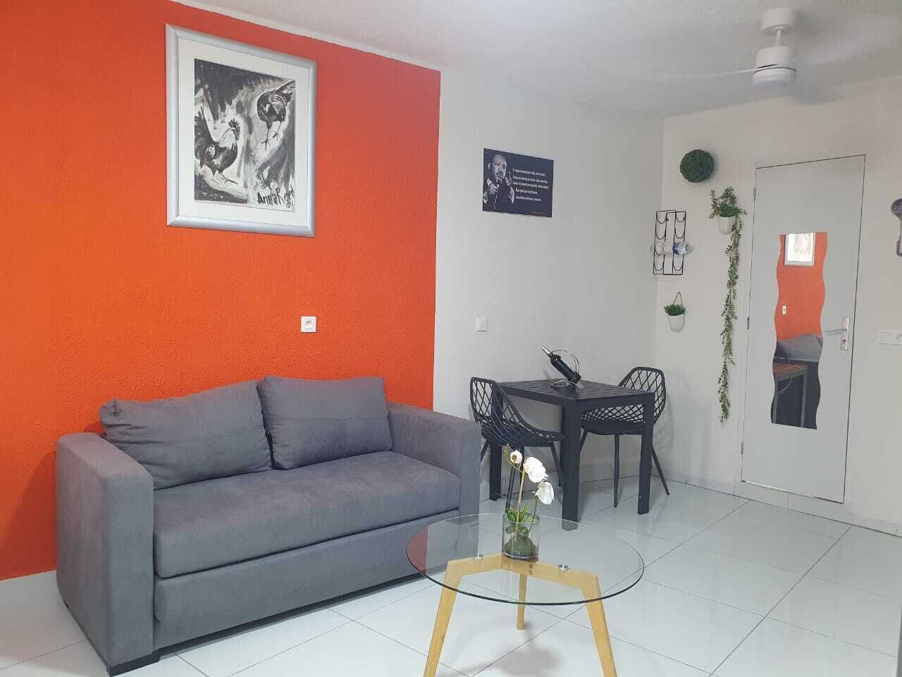 Una sala de estar con un sofá gris, una mesa de centro de vidrio, una mesa de comedor con sillas y una puerta con un espejo.