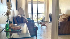 A living room with a glass-top dining table, wicker furniture, and a view of the outdoors through large windows. - Cozy condo with a view of famous Kamaole Beach II (Kihei)