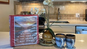 A kitchen with a microwave, cabinets, a sink, a decorative orchid, a book, and two mugs. - Cozy condo with a view of famous Kamaole Beach II (Kihei)