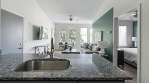 A modern kitchen with a granite countertop and a built-in sink, a dining area with a table and chairs, a living room with a sofa and a TV, and a bedroom with a bed and a nightstand. - Landing | Beautiful 1BD, Pool, Gym (Nashville)