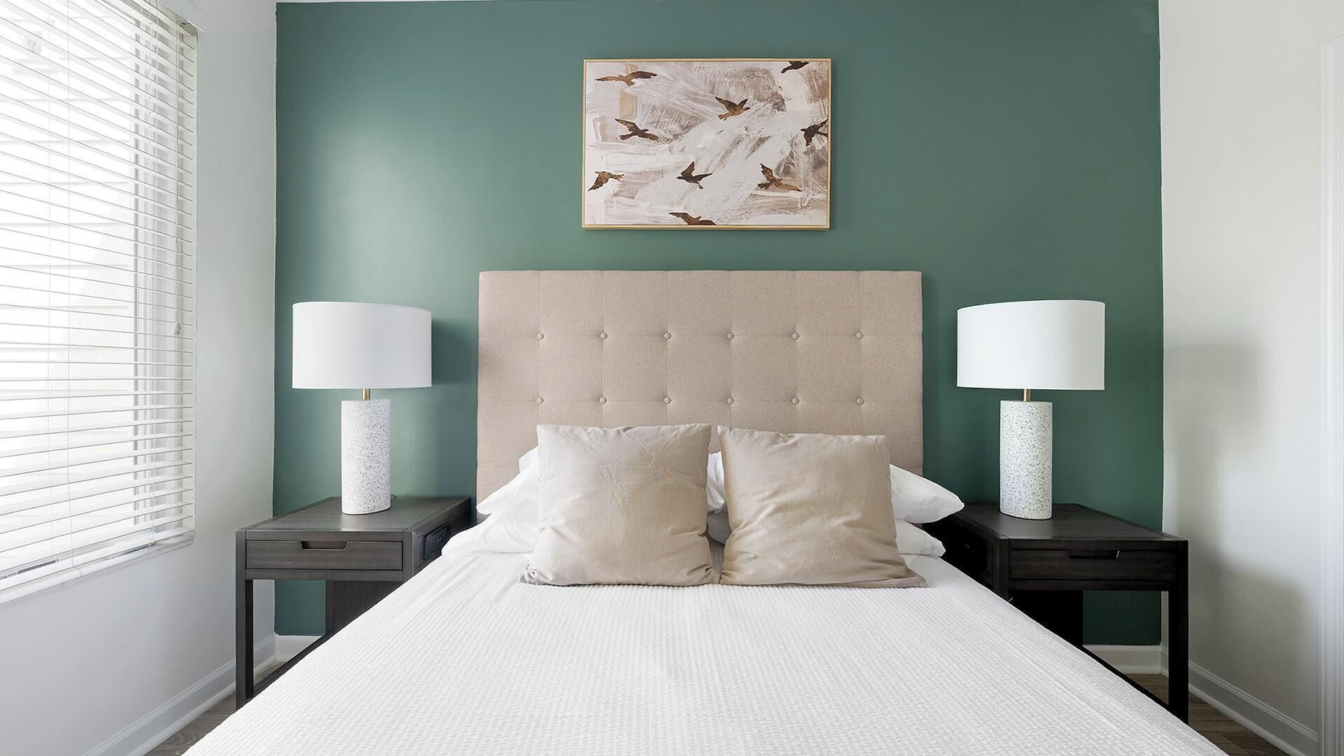 Un dormitorio con cabecera capitonada, dos mesitas de noche con lámparas, una cama con ropa de cama blanca y una obra de arte en la pared con pájaros.